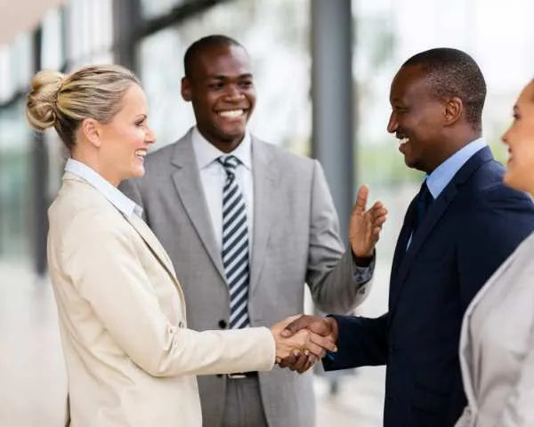 Two people shaking hands in an office walkway while two colleagues stand alongside, mid-conversation.